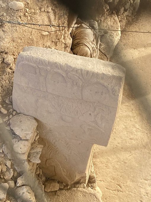 The image features an old stone carving, possibly a monument or a statue, situated on a dirt road surrounded by rocks. The carving appears to be weathered and worn, suggesting that it has been exposed to the elements for quite some time. In the background, there is a wire fence running along the side of the road, adding to the rustic atmosphere of the scene.
