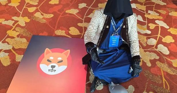 A person in a blue and silver costume with a black face covering sits on a patterned carpet next to a Shiba Inu dog logo sign.