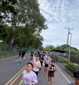 A group of people participate in a road race, running down a tree-lined street on what appears to be a sunny morning, with power lines visible overhead and a runner wearing a bib with '24' in the foreground.