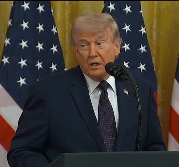 The image features a man wearing a suit and tie standing at a podium with his mouth open while speaking into a microphone, suggesting he is delivering a speech or addressing an audience. The backdrop includes American flags, indicating that this event might be taking place in the United States. The text "NEW: Donald Trump said the U.S. must protect its leadership in prediction markets and crypto, warning that other countries are trying to overtake America as the “Bitcoin capital of the world." is displayed on the image, which seems to be related to a political speech or statement by the President.