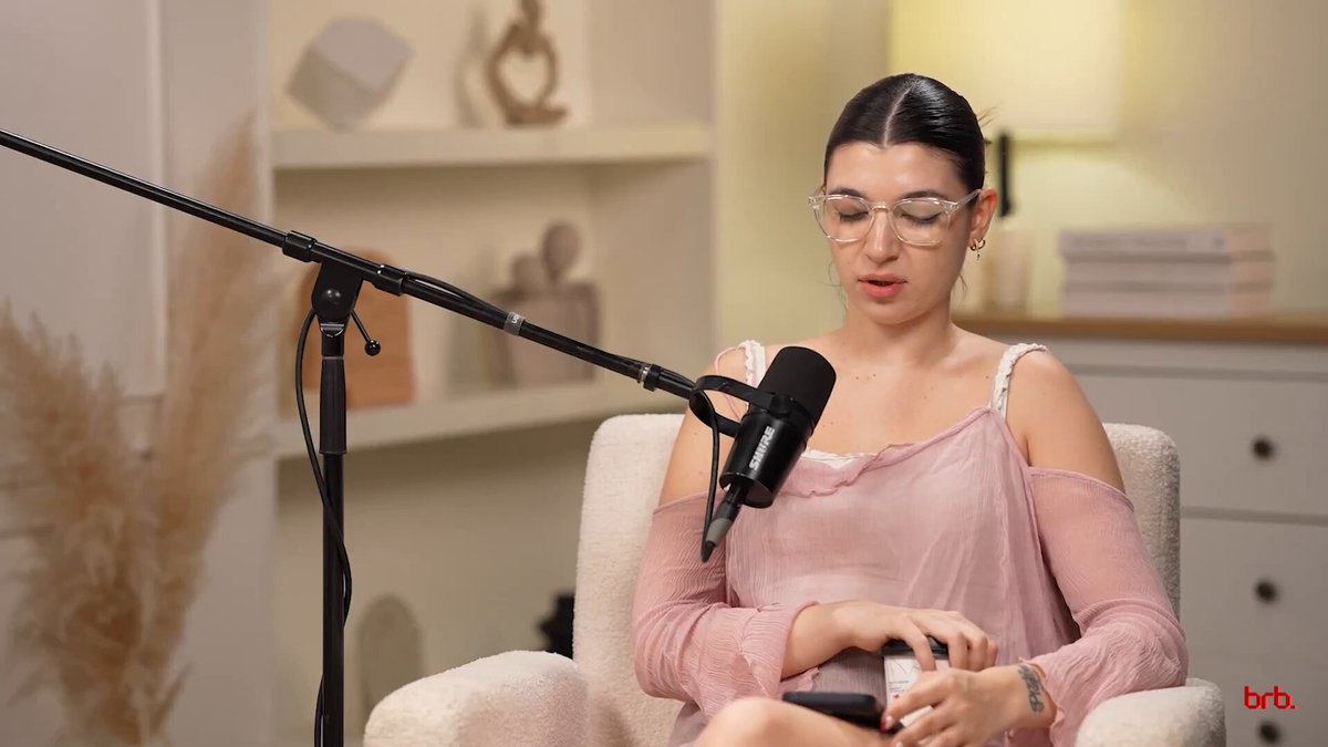 The image features a woman sitting in an armchair with her legs crossed while wearing glasses and a pink top. She appears to be talking into a microphone, possibly discussing Bitcoin or related topics. The background includes a bookshelf filled with various items such as vases and a clock.