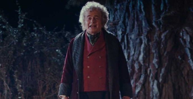 An older man with wild gray hair, wearing a red vest and dark coat, stands in a dark forest setting, appearing to speak.