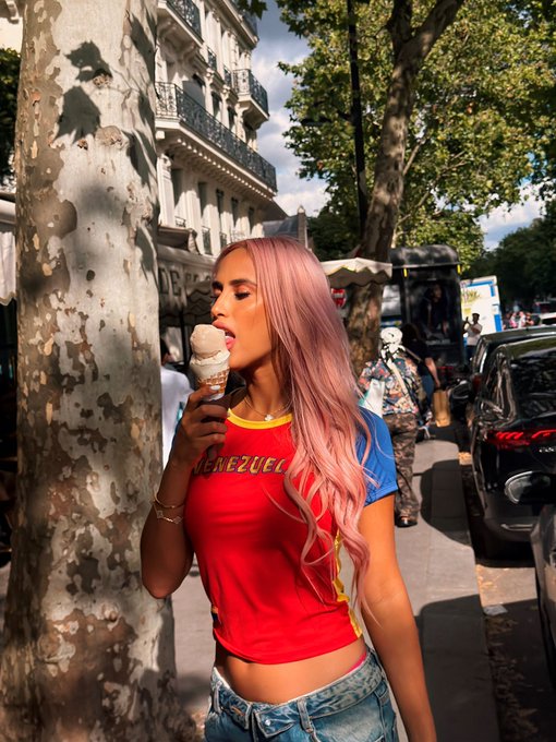 A young woman with long pink hair enjoys an ice cream cone on a sunlit Parisian street, wearing a red and blue top reading "CHEZUELA" with yellow trim. The relaxed summer vibe and ice cream treat tie into the "SOLANA SUMMER" post, evoking a carefree, sunny season.