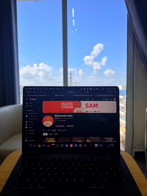 A laptop on a desk displays a YouTube channel called 'Blockchain Sam' featuring a banner with the text 'BLOCK CHAIN SAM' and 'أكاديمية سلسلة الكتل' (Blockchain Academy). The scene is framed by a window with blue curtains showing a bright sky and clouds over water.