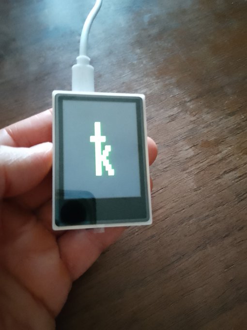 A hand holding a small white hardware wallet device connected to a charging cable, with a pixelated 'k' logo displayed on its screen.