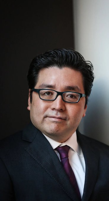A man in a dark suit, white shirt, and purple tie with black-rimmed glasses poses for a professional portrait against a dark background. The image likely represents one of the prominent investors mentioned in the post, such as Michael Saylor or Tom Lee, who are noted for holding massive unrealized losses without selling their crypto positions like $MSTR.