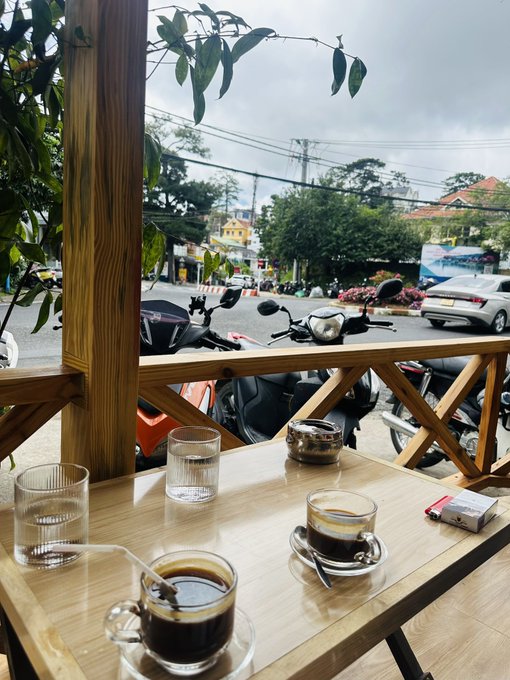 A view from a wooden café table with two glasses of Vietnamese coffee, looking out over a street scene with parked motorbikes and a white car on an overcast day.