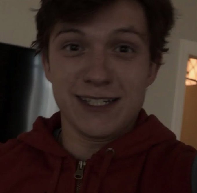 A young man wearing a red hoodie smiles at the camera in what appears to be a selfie taken indoors, with a doorway visible in the background. The image humorously represents the "Average $HBAR Holder" meme comparing a cheerful 2024 investor to a presumably less cheerful 2026 version, capturing the optimistic expression of someone still hopeful about their Hedera holdings.