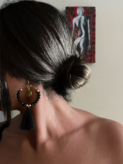 A close-up view from behind shows a woman with dark hair in a bun, wearing a black beaded earring with a coin charm and tassel, while a red and black painting hangs on the wall in the background.