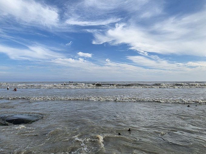 A serene beach landscape showing waves rolling onto the shore under a bright blue sky with scattered white clouds, with a few distant figures in the water.