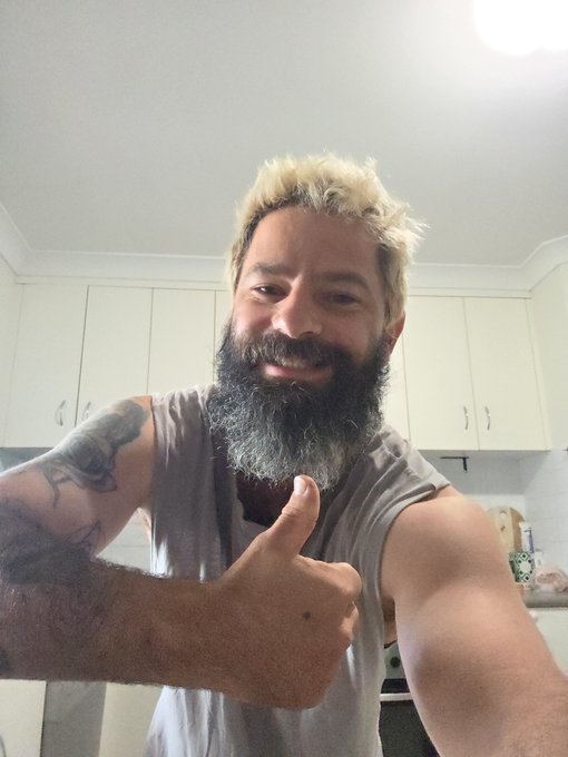 A man with blonde hair and a beard gives a thumbs-up while taking a selfie in what appears to be a kitchen, with white cabinets visible in the background.