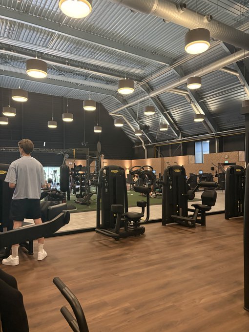 A spacious fitness center with wood flooring, industrial ceiling, and pendant lights shows a person in a gray t-shirt and dark shorts standing among various weight machines and workout equipment.