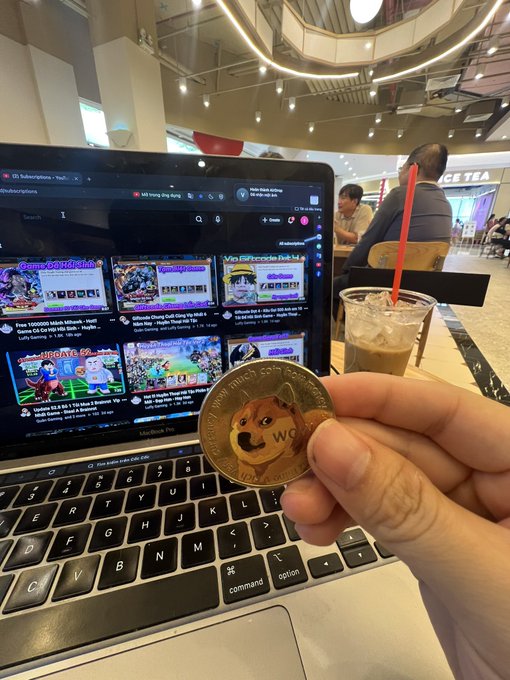The image shows a person holding up an old coin with a cartoon dog on it, which has been posted online for sale. The coin appears to be worth 100 dollars. In the background of the photo, there are other people sitting at tables or standing around in what seems to be a cafe setting.