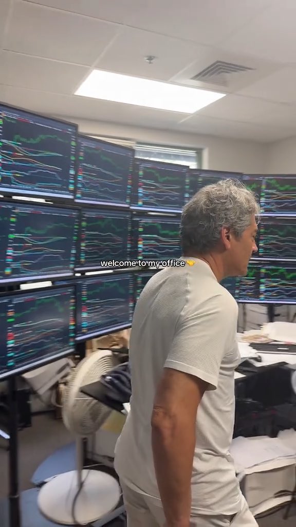 A man stands in his office surrounded by multiple monitors displaying trading charts, with the caption 'welcome to my office 👉' overlaid.