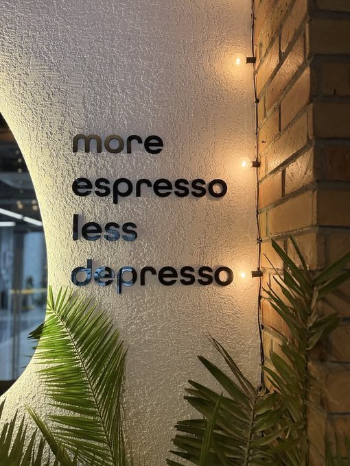 A textured wall features the playful text 'more espresso less depresso' in black lettering, illuminated by string lights next to a brick column and tropical plants.