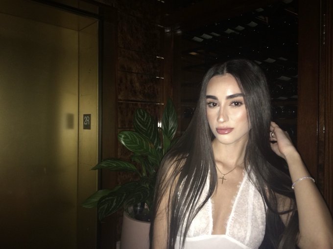A young woman with long dark hair poses indoors wearing a white lace top and a cross necklace, with a plant visible behind her.