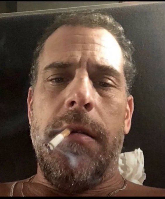 A close-up portrait of a bearded man with graying hair looking directly at the camera while smoking a cigarette.