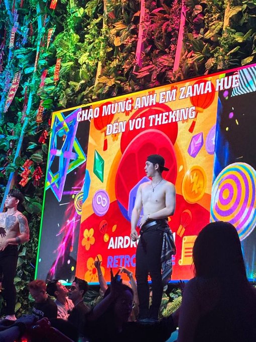 A shirtless man performs on stage at a venue with a colorful screen behind him displaying Vietnamese text including 'CHAO MUNG ANH EM ZAMA HUE DEN VOI THEKING' and 'AIRDROP RETRO'.