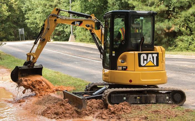 The image features a yellow Cat excavator with its bucket raised, digging into a muddy area near a road. The excavator appears to be working hard as it digs through the dirt. In the background, there are trees visible, adding to the outdoor setting of the scene. The photo is accompanied by a caption that reads "Getting pretty close to a 5X on CAT in the past 4 years Think I might buy one of these to celebrate". This suggests that the excavator has been used for some time and may have had an impact on its owner or others who appreciate the history behind it.