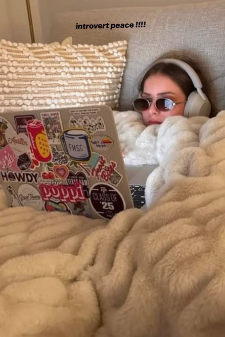 A woman wearing sunglasses and headphones works on a laptop covered in stickers while wrapped in a fuzzy white blanket, with the caption 'introvert peace !!!!' above her.