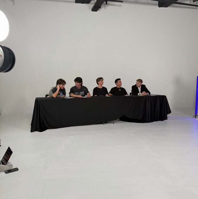 Five young men sit behind a long black-draped table in a bright white photo studio with professional lighting equipment visible, with one wearing a "SATURDAY" t-shirt, gathered as they prepare for their upcoming content series launch.
