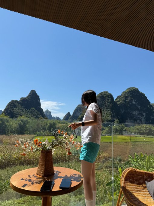 A woman stands on a balcony overlooking a stunning landscape of lush green karst mountains, with a wooden table in the foreground holding a vase of orange flowers and two smartphones. The serene view of rolling green peaks and vibrant vegetation perfectly captures the post's reference to "bullish green," suggesting a market outlook as uplifting and promising as the verdant vista.