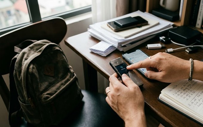 A person sits at a wooden desk near a window, holding a smartphone in one hand while tapping another phone with the other, surrounded by stacks of papers, a backpack, and an open notebook with handwritten notes.