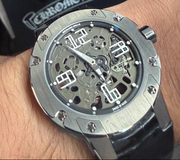 A close-up of a skeleton-dial luxury watch with a stainless steel octagonal case and black leather strap worn on the wrist, with 'CHRONO' partially visible in the background.