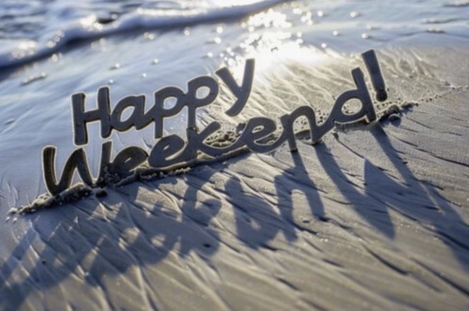 The text "Happy Weekend!" written in a wavy, textured style on wet sand at a beach, with sunlight glistening on the water in the background.