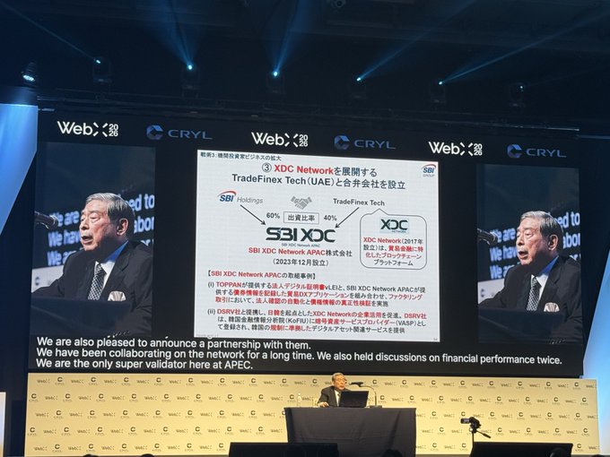 An SBI executive presents at the WebX conference, with a slide announcing the joint venture between SBI Holdings and TradeFinex Tech (UAE) to establish XDC Network APAC, with SBI holding a 60% stake and TradeFinex Tech 40% (SBI XDC Network APAC株式会社, established December 2023). The bottom text reads, "We are also pleased to announce a partnership with them. We have been collaborating on the network for a long time. We also held discussions on financial performance twice. We are the only super validator here at APEC."