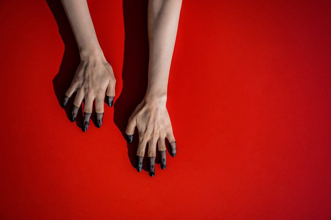Two pale hands with black painted fingertips resting against a bold red background.