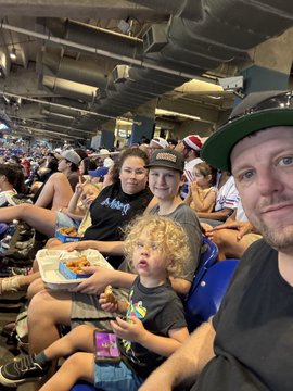 A group including children sit in stadium seats enjoying food and drinks, with one child holding a clapper and another resting a tray of snacks on their lap. The caption highlights the moment as the essence of the experience, showing "Kups holders" enjoying the event alongside sponsors like Raposa Coffee Co.