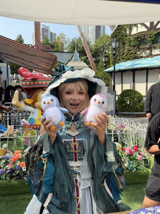 A smiling person in an elaborate teal and white ruffled costume holds up two plush bird toys at what appears to be a theme park.