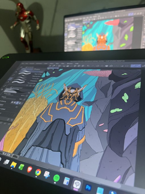 A drawing tablet displaying a digital illustration of a fantasy character in a dark hooded robe with an animal skull mask, with Clip Studio Paint interface visible and a second monitor showing similar artwork in the background, alongside a small Iron Man figurine.