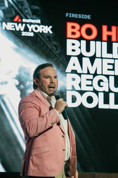 A man in a pink blazer speaks into a microphone at a Summit New York 2025 event, with a backdrop reading 'FIRESIDE' and partially visible text 'BUILD AMERICA REGU[DOLLAR]'.