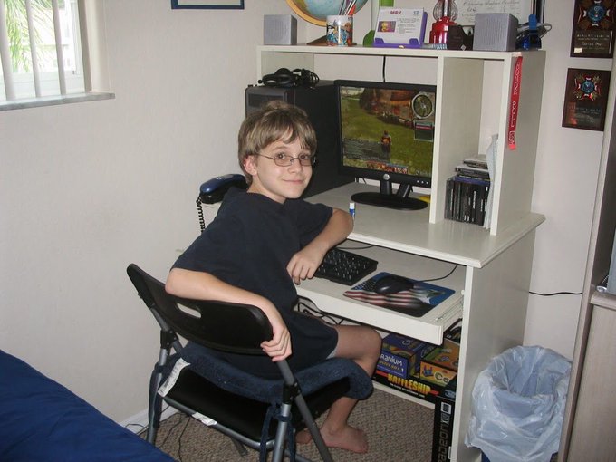 A young boy with glasses sitting at a computer desk, smiling at the camera, with a monitor displaying a video game and various items including board games and a trash can nearby.