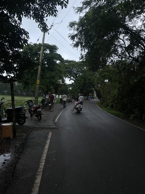 A street scene with motorbikes and pedestrians under a cloudy sky, showing parked motorcycles along a wet road lined with trees.