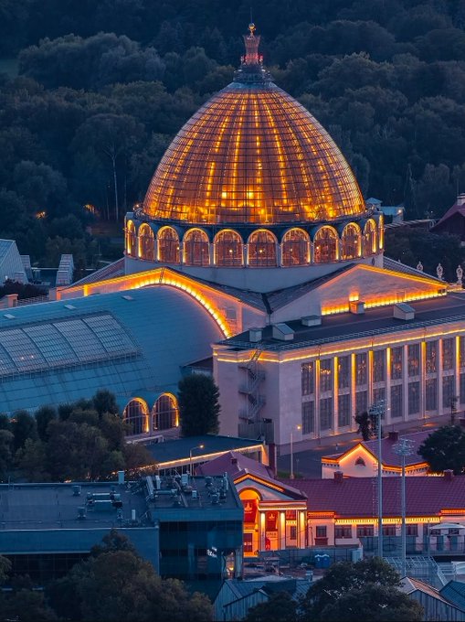 A nighttime aerial view of an illuminated glass dome building, likely a conservatory or pavilion, glowing with warm orange lights against a dark forest backdrop.