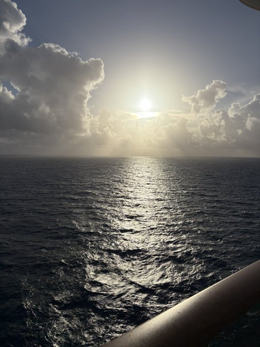 A scenic photograph taken from a ship of the sun shining through scattered clouds, casting a silver reflection across the vast dark ocean.