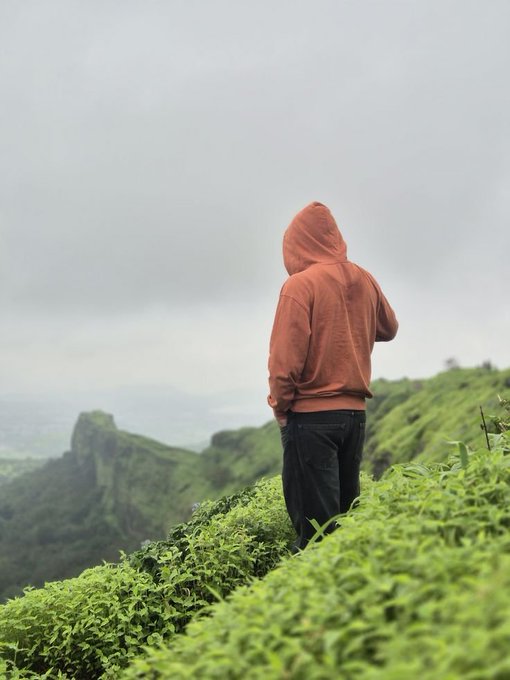 A person in an orange hoodie stands alone on a lush green hilltop, gazing out at a misty mountainous landscape. The image reflects the post's message of patience and steady perseverance—much like the solitary figure taking in the expansive view, the post encourages $ANSEM holders to embrace slow, consistent growth over temporary hype.