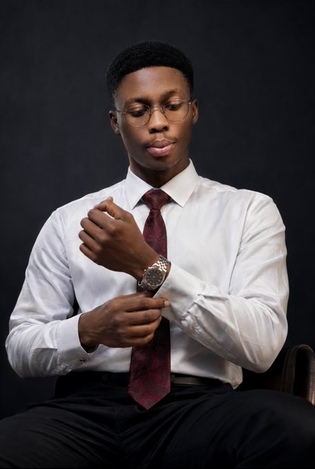 A young man wearing glasses, a white dress shirt, and a burgundy patterned tie, adjusting his cuffs while wearing a luxury watch, photographed against a dark background.