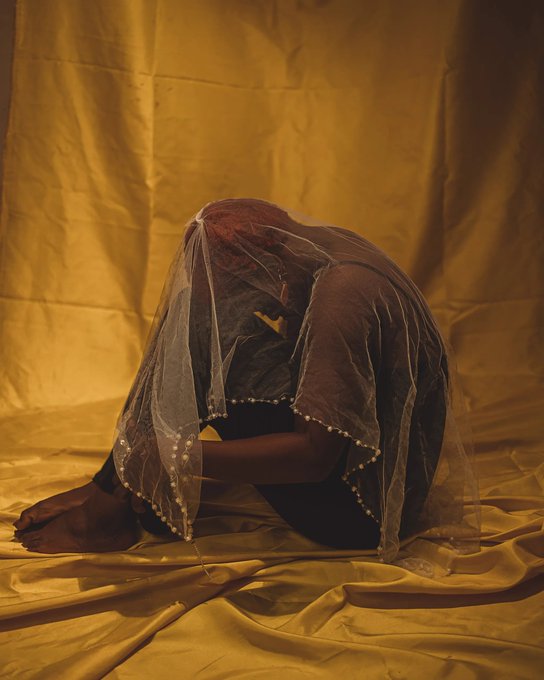 A person draped in a sheer, embroidered veil sits curled up against a golden-yellow fabric backdrop in warm lighting.