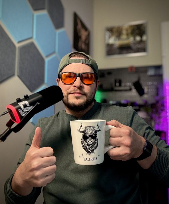 A bearded man wearing a green cap and orange-tinted sunglasses gives a thumbs-up while holding a white mug featuring a Highland cow illustration and the word 'TAURUS', seated in front of a microphone.