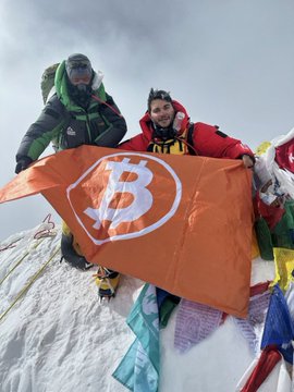 In the image, two hikers are standing on top of a mountain, holding an orange Bitcoin-themed flag. They appear to be celebrating their achievement or commemorating their journey up the mountain. The flag has a white "B" symbolizing Bitcoin on its surface and is held aloft by the hikers as they pose for the photo.