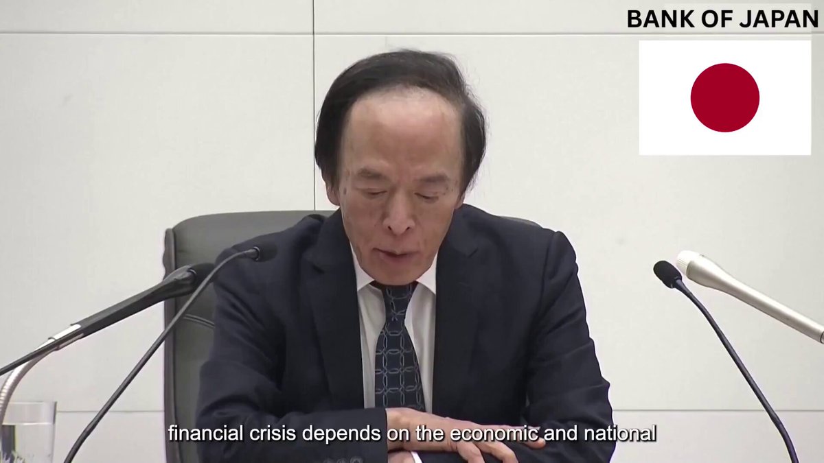 The image shows a man sitting at a desk, possibly in front of microphones or at a press conference. He appears focused and serious while wearing a suit and tie, suggesting that he is involved in some form of public speaking event or addressing an important issue. The background features elements related to the Bank of Japan, such as a red flag symbolizing national significance. There are also text present on this image, though their content is not explicitly clear from the provided details.