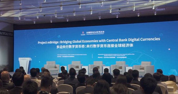 A conference room shows an audience seated in front of a large blue screen displaying 'Project mbridge: Bridging Global Economies with Central Bank Digital Currencies' in English and Chinese.