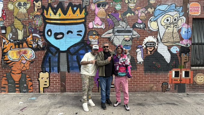 Three people posing together in front of a large, colorful mural featuring various cartoon-like characters painted on a brick wall, with a sign reading 'Pedro King Streetwear' in the top right.