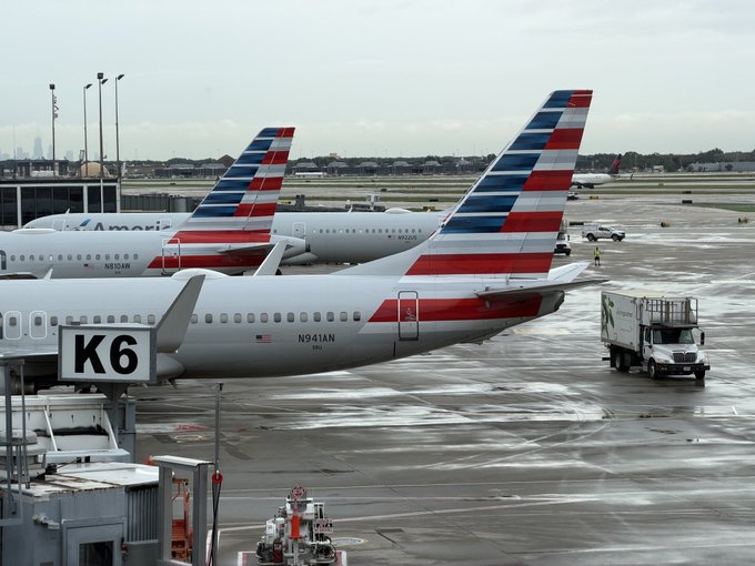 Several American Airlines planes, with visible registration numbers N941AN and N101NN, are parked at gate K6 of an airport, with a catering truck servicing one aircraft. The image evokes the "heading to New York" travel theme from the post, setting the scene for the speaker's journey to the UN Headquarters to discuss AI and blockchain.