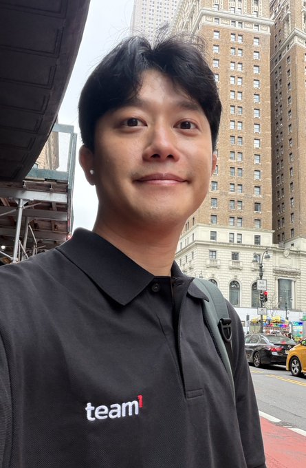 A selfie of a man wearing a black polo shirt with 'team1' printed on it, standing on a city street with tall brick buildings and a yellow taxi visible in the background.