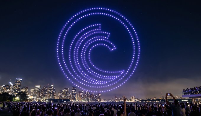 A nighttime drone light show forms a large spiral pattern of purple lights above a city skyline, while a crowd watches and photographs the spectacle from the waterfront.